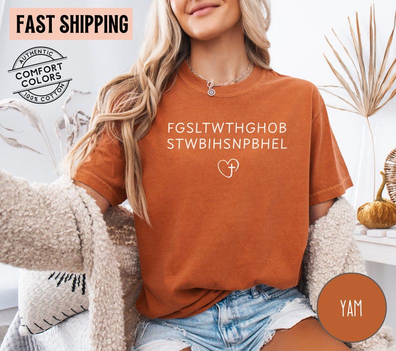 May include: A burnt orange t-shirt with white text and a heart with a cross graphic. The shirt has the words "AUTHENTIC COMFORT COLORS 100% COTTON" printed on it. The shirt is made of a soft material.