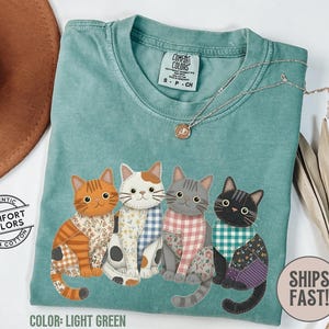 May include: Light green Comfort Colors t-shirt featuring a colorful graphic of four cats with patterned bandanas and patchwork details. The shirt has a crew neck and short sleeves. The text "COMFORT COLORS" is visible on the tag.