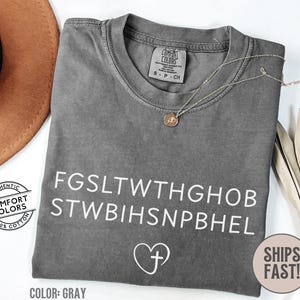 May include: Gray Comfort Colors t-shirt with white text and a heart with a cross graphic. The shirt has a crew neck and short sleeves. A silver necklace with a pendant is on the shirt. The shirt is made of 100% cotton.