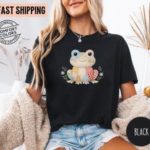 May include: Black t-shirt featuring a colorful patchwork frog design. The frog has a smiling face and is surrounded by floral accents. The shirt is made of 100% cotton and has the words "Authentic Comfort Colors" printed on it.