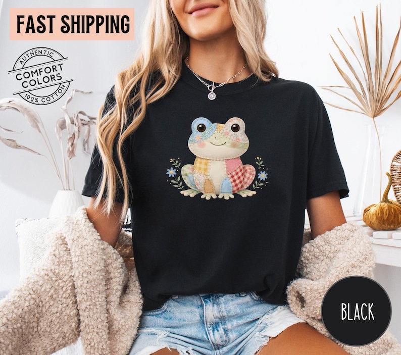 May include: Black t-shirt featuring a colorful patchwork frog design. The frog has a smiling face and is surrounded by floral accents. The shirt is made of 100% cotton and has the words "Authentic Comfort Colors" printed on it.