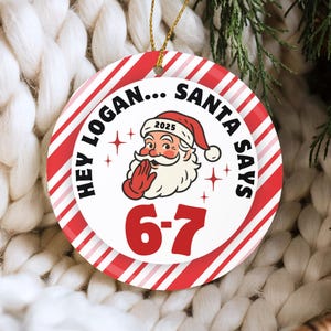 May include: A round Christmas ornament with a red and white striped border. The center features Santa Claus with the text "HEY LOGAN... SANTA SAYS" and the numbers "6-7". The ornament is hanging by a gold string.