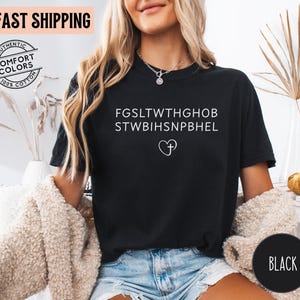May include: Black t-shirt with white text and a heart-shaped design with a cross. The shirt has the words "AUTHENTIC COMFORT COLORS 100% COTTON" printed on it. The words "FAST SHIPPING" are printed on the top left of the image.