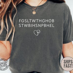 May include: A dark gray Comfort Colors t-shirt with white text and a heart-shaped cross graphic. The text includes two lines of seemingly random letters. A circular logo with the words "Comfort Colors" and "Authentic 100% Cotton" is visible.