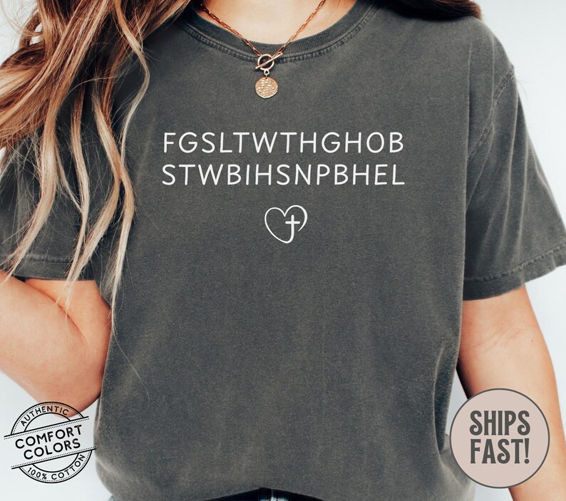 May include: A dark gray Comfort Colors t-shirt with white text and a heart-shaped cross graphic. The text includes two lines of seemingly random letters. A circular logo with the words "Comfort Colors" and "Authentic 100% Cotton" is visible.