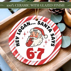 May include: A round ceramic Christmas ornament with a red and white striped border. The ornament features Santa Claus with the text "HEY LOGAN... SANTA SAYS 6-7" and the year 2025. The ornament is on a wooden tray.