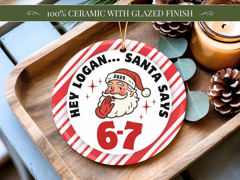 May include: A round ceramic Christmas ornament with a red and white striped border. The ornament features Santa Claus with the text "HEY LOGAN... SANTA SAYS 6-7" and the year 2025. The ornament is on a wooden tray.