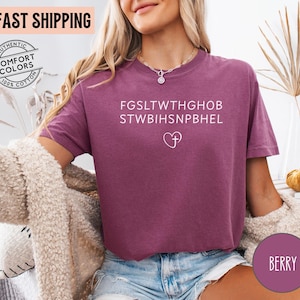 May include: Berry-colored t-shirt with white text and a heart-shaped cross design. The shirt has a relaxed fit and is made of a soft material. The shirt also has the text "FAST SHIPPING" and "AUTHENTIC COMFORT COLORS 100% COTTON" on the top left.