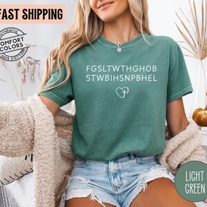 May include: Light green t-shirt with white text and a heart-shaped cross design. The shirt has the words "Authentic Comfort Colors 100% Cotton" printed on it. The words "Fast Shipping" are printed on the top left.