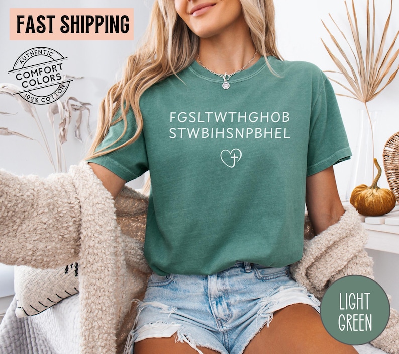 May include: Light green t-shirt with white text and a heart-shaped cross design. The shirt has the words "Authentic Comfort Colors 100% Cotton" printed on it. The words "Fast Shipping" are printed on the top left.