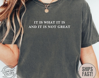 May include: A dark gray t-shirt with the text "IT IS WHAT IT IS AND IT IS NOT GREAT" printed in white.