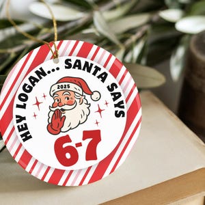 May include: A round Christmas ornament with a red and white striped border. The center features Santa Claus with the text "HEY LOGAN... SANTA SAYS 6-7 2025". A gold string is attached for hanging.