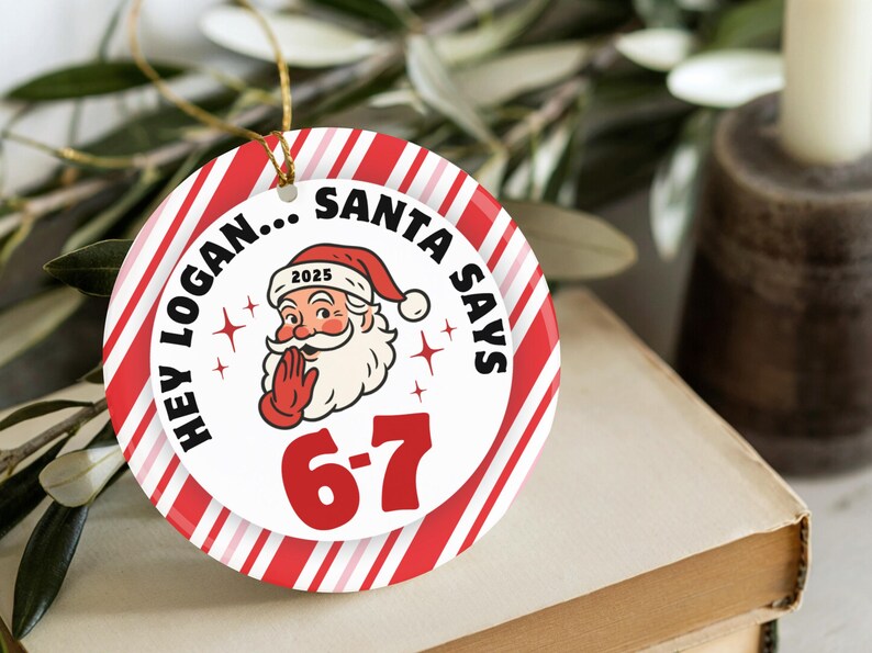 May include: A round Christmas ornament with a red and white striped border. The center features Santa Claus with the text "HEY LOGAN... SANTA SAYS 6-7 2025". A gold string is attached for hanging.