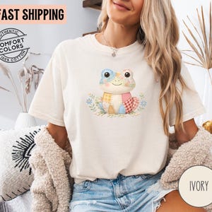 May include: Ivory-colored t-shirt featuring a colorful patchwork frog design. The frog has a smiling face and is surrounded by floral accents. The shirt also has the text "FAST SHIPPING" and "AUTHENTIC COMFORT COLORS 100% COTTON".
