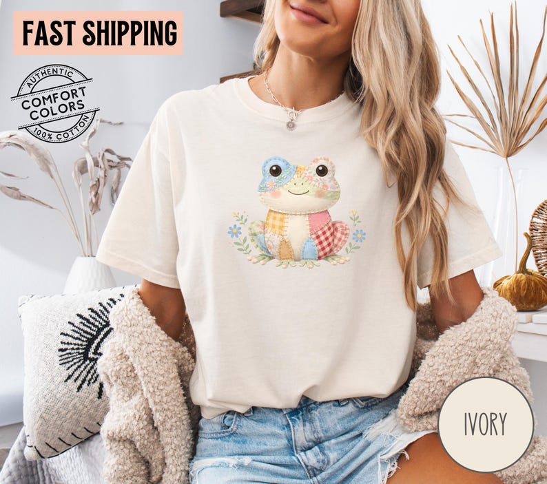 May include: Ivory-colored t-shirt featuring a colorful patchwork frog design. The frog has a smiling face and is surrounded by floral accents. The shirt also has the text "FAST SHIPPING" and "AUTHENTIC COMFORT COLORS 100% COTTON".