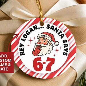 May include: A round Christmas ornament with a red and white striped border. The ornament features Santa Claus with the text "HEY LOGAN... SANTA SAYS" and "6-7". The year "2025" is on Santa's hat. A gold string is attached.