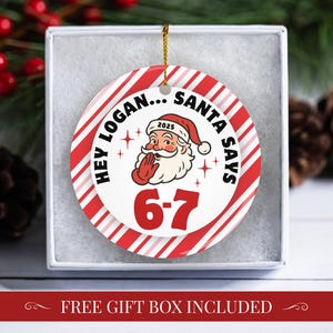 May include: A round Christmas ornament with a red and white striped border. The ornament features Santa Claus with the text "HEY LOGAN... SANTA SAYS 6-7 2025". The ornament is in a white gift box.