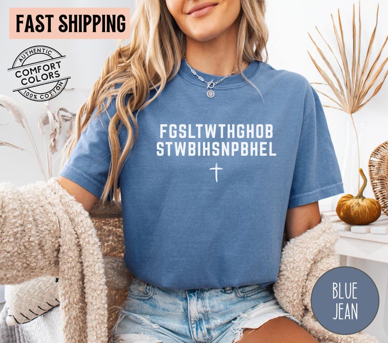 Puede incluir: Una camiseta azul de manga corta con texto blanco que dice FGSLTWTHGHOB STWBIHSNPBHEL y un peque&ntilde;o s&iacute;mbolo de cruz blanca centrado debajo del texto. La prenda tiene cuello redondo y un corte holgado.