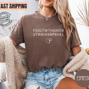 May include: Espresso-colored t-shirt with white text and a heart-shaped cross graphic. The shirt is made of 100% cotton and features the words "Authentic Comfort Colors". The shirt is paired with denim shorts.