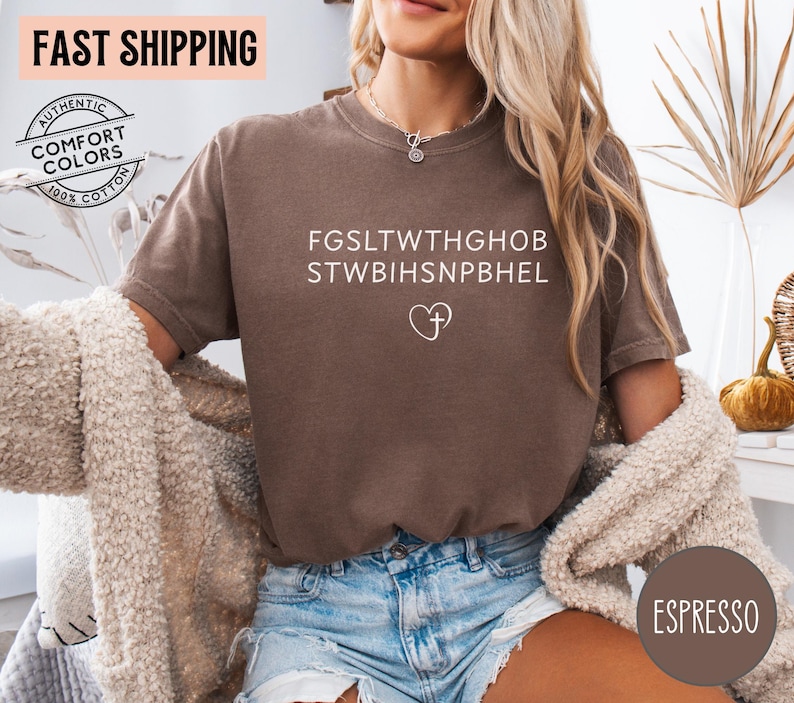 May include: Espresso-colored t-shirt with white text and a heart-shaped cross graphic. The shirt is made of 100% cotton and features the words "Authentic Comfort Colors". The shirt is paired with denim shorts.