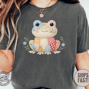 May include: Dark gray Comfort Colors t-shirt featuring a patchwork frog design. The frog has a variety of colorful fabric patches and is surrounded by small blue flowers and green leaves. The shirt also has the text "Comfort Colors" and "Ships Fast!"