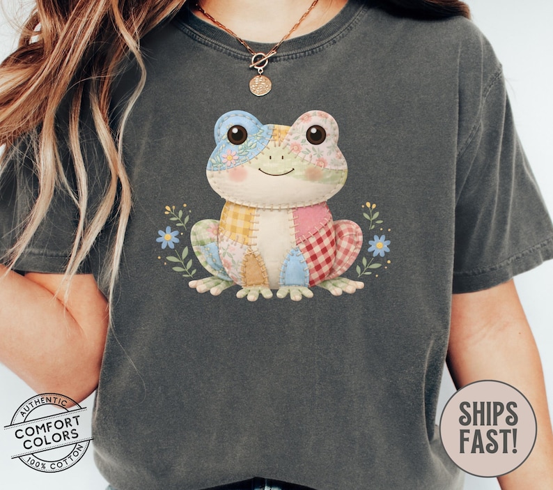 May include: Dark gray Comfort Colors t-shirt featuring a patchwork frog design. The frog has a variety of colorful fabric patches and is surrounded by small blue flowers and green leaves. The shirt also has the text "Comfort Colors" and "Ships Fast!"
