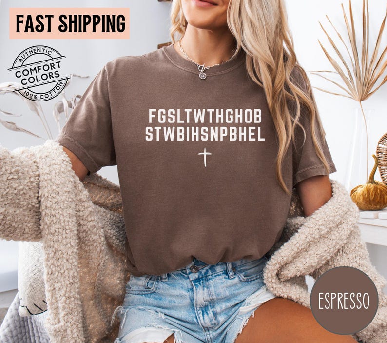 Puede incluir: Camiseta color espresso con texto blanco y cruz. La camiseta tiene cuello redondo y mangas cortas. El texto de la camiseta es en negrita, sin serifa. La camiseta est&aacute; hecha de un material suave y c&oacute;modo.