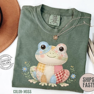May include: Moss green t-shirt featuring a patchwork frog design. The frog has blue, yellow, pink, and red checkered patterns. The shirt has a crew neck and short sleeves. The text "SHIPS FAST!" is on the bottom right.