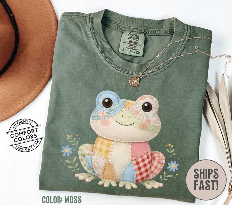 May include: Moss green t-shirt featuring a patchwork frog design. The frog has blue, yellow, pink, and red checkered patterns. The shirt has a crew neck and short sleeves. The text "SHIPS FAST!" is on the bottom right.