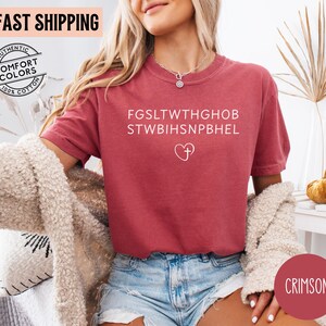 May include: Crimson-colored t-shirt with white text and a heart-and-cross graphic. The shirt features the text "FGS...HEL" and is made of 100% cotton. The model is wearing a necklace and blue denim shorts.