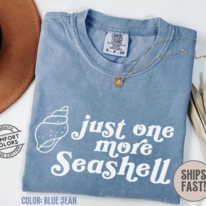 May include: Blue jean Comfort Colors t-shirt with the text "just one more seashell" in white. A seashell graphic is on the left. A gold necklace with a pendant is on the shirt. The shirt has a small tag that says "Comfort Colors".