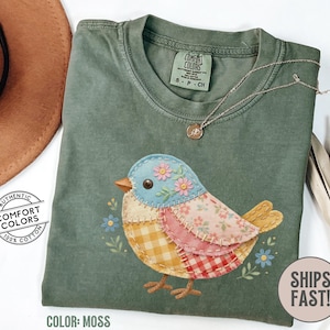 May include: Moss green Comfort Colors t-shirt with a colorful patchwork bird design. The bird features blue, pink, yellow, and red fabrics with floral accents. The shirt has a crew neck and short sleeves. The text "SHIPS FAST!" is visible.