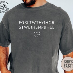 May include: Dark gray t-shirt with white text and a heart-shaped cross design. The shirt features the phrases "FGSLTWTHGHOB" and "STWBIHSNPBEL" above the heart and cross symbol. The shirt is made of 100% cotton.