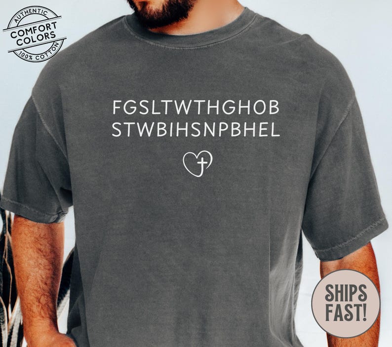 May include: Dark gray t-shirt with white text and a heart-shaped cross design. The shirt features the phrases "FGSLTWTHGHOB" and "STWBIHSNPBEL" above the heart and cross symbol. The shirt is made of 100% cotton.