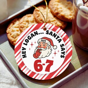 May include: A round Christmas ornament with red and white stripes and the text "Hey Logan... Santa Says 6-7". The ornament features a Santa Claus illustration and the year 2025. The ornament is on a tray with cookies and a mug of hot chocolate.