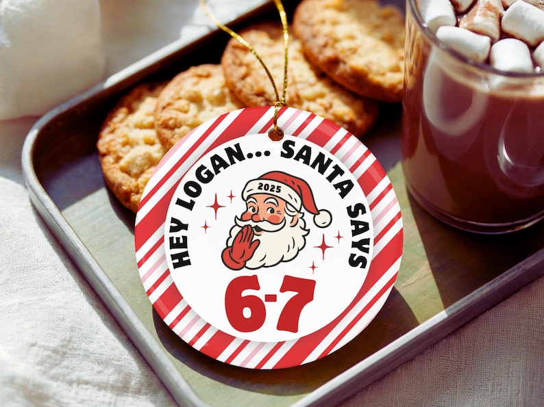 May include: A round Christmas ornament with red and white stripes and the text "Hey Logan... Santa Says 6-7". The ornament features a Santa Claus illustration and the year 2025. The ornament is on a tray with cookies and a mug of hot chocolate.