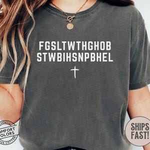 Puede incluir: Camiseta gris oscuro con texto blanco y una cruz. El texto est&aacute; dispuesto en dos l&iacute;neas de may&uacute;sculas. La camiseta tiene un corte holgado y mangas cortas. Hecha de 100% algod&oacute;n.