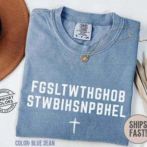 Puede incluir: Camiseta Comfort Colors azul vaquero con texto blanco y un gr&aacute;fico de cruz. La camiseta tiene cuello redondo y mangas cortas. Un collar dorado con un colgante est&aacute; sobre la camiseta. La camiseta es 100% algod&oacute;n.