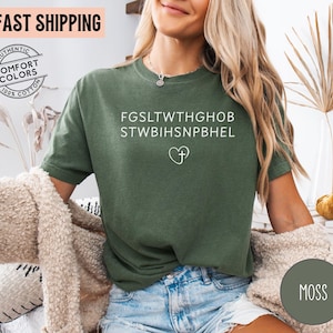 May include: Moss green t-shirt with white text and a heart-shaped cross design. The shirt is made of 100% cotton and features the words "Authentic Comfort Colors". The shirt also has the words "FAST SHIPPING" on the top left.