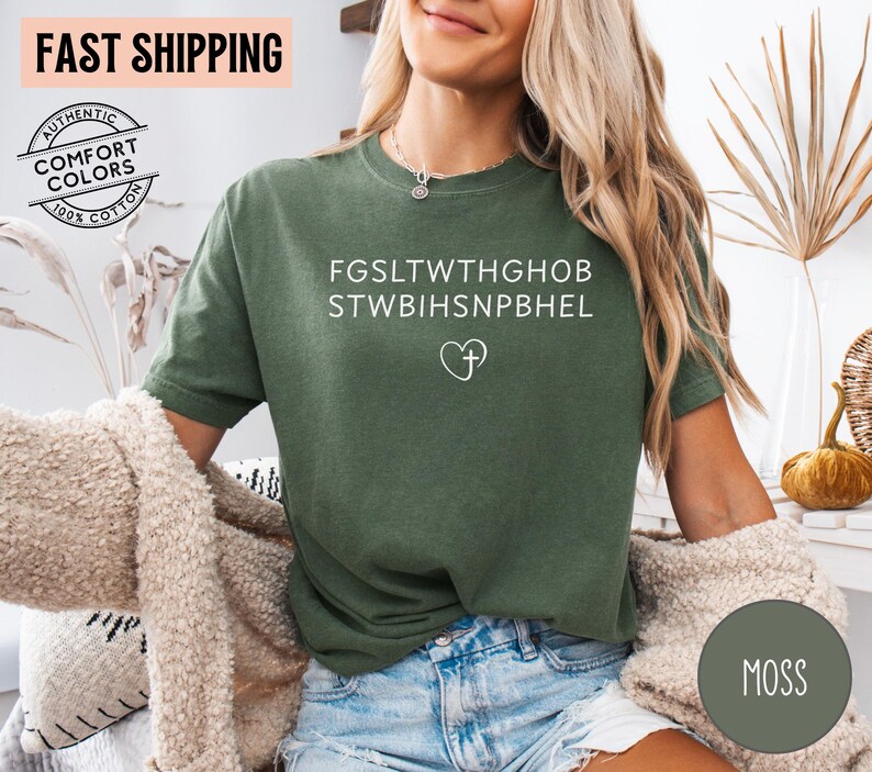 May include: Moss green t-shirt with white text and a heart-shaped cross design. The shirt is made of 100% cotton and features the words "Authentic Comfort Colors". The shirt also has the words "FAST SHIPPING" on the top left.