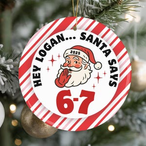 May include: Round Christmas ornament with a red and white striped border. The ornament features the text "HEY LOGAN... SANTA SAYS" around a Santa Claus graphic. The numbers "6-7" are in red below Santa.