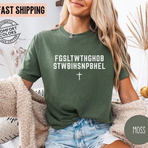 Puede incluir: Camiseta verde musgo con texto blanco y cruz. La camiseta presenta el texto "FGSLTWTHGHOB STWBIHSNPBHEL". Hecha de 100% algod&oacute;n, tiene un aspecto vintage.