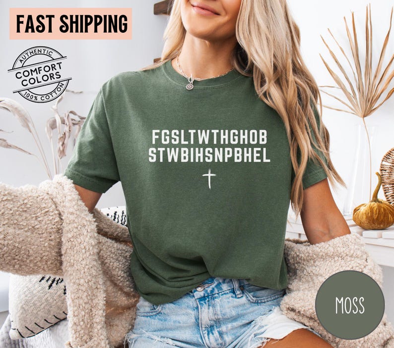 Puede incluir: Camiseta verde musgo con texto blanco y cruz. La camiseta presenta el texto "FGSLTWTHGHOB STWBIHSNPBHEL". Hecha de 100% algod&oacute;n, tiene un aspecto vintage.