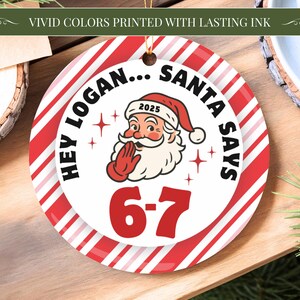 May include: A round Christmas ornament with a red and white striped border. The center features Santa Claus with the text "HEY LOGAN... SANTA SAYS 6-7" and the year 2025. The ornament is hanging by a gold string.