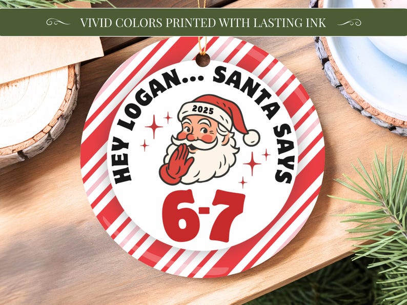 May include: A round Christmas ornament with a red and white striped border. The center features Santa Claus with the text "HEY LOGAN... SANTA SAYS 6-7" and the year 2025. The ornament is hanging by a gold string.