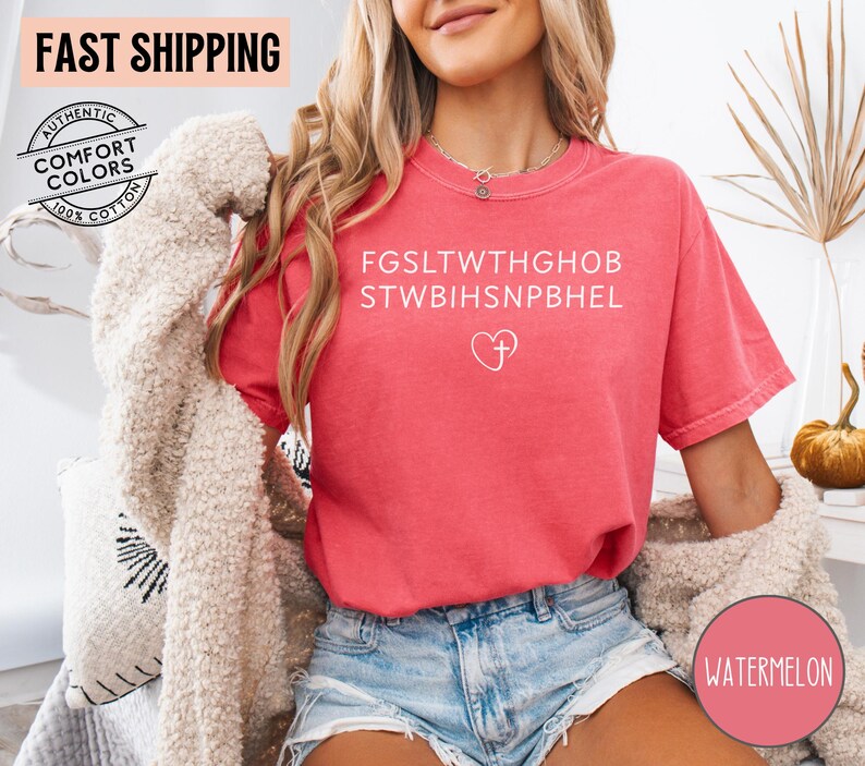 May include: A watermelon-colored t-shirt with white text and a heart with a cross graphic. The shirt has short sleeves and a relaxed fit. The words "FAST SHIPPING" and "AUTHENTIC COMFORT COLORS 100% COTTON" are also visible.