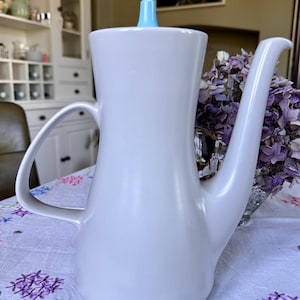 May include: A white ceramic teapot with a light blue lid and a curved spout. The teapot is sitting on a white tablecloth with a floral pattern.