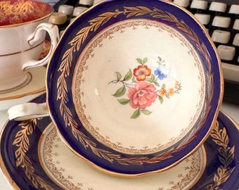 May include: A cream-coloured teacup and saucer set with a navy blue border and gold leaf detailing. The teacup features a floral design in the centre. The set is placed in front of a vintage typewriter, evoking a sense of nostalgia.