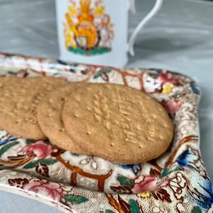 May include: A white ceramic tray with a floral pattern in shades of pink, green, blue, and brown. The tray has a scalloped edge and features a raised design. Three round, brown biscuits are on the tray.