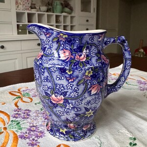以下が含まれることがあります： ピンクと黄色の花柄の青いセラミックピッチャー。ピッチャーは白い縁と湾曲したハンドルが付いています。背景には白いキャビネットと、花の刺繍が施されたテーブルクロスのあるテーブルが含まれています。